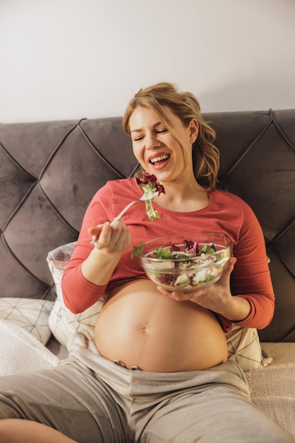 Happy pregnant woman eating fresh vegetable salad while sitting on the bed in her bedroom.