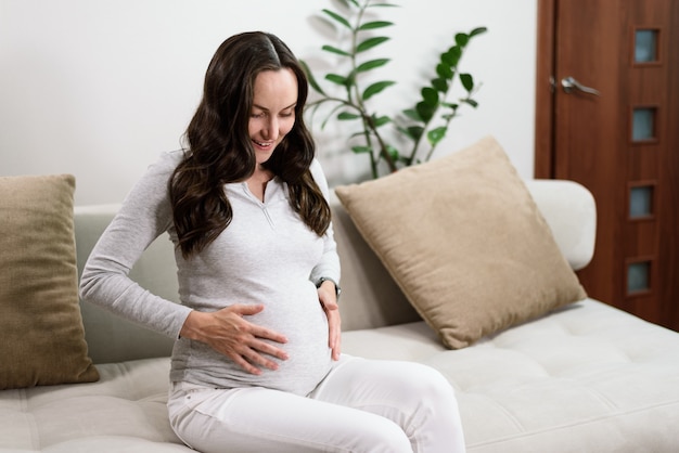 Happy pregnant woman in grey long sleeved t-shirt with hand on belly sitting on sofa , happy pregnancy concept, fetal motions