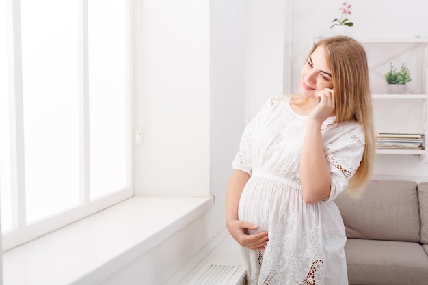 Happy pregnant woman talking on her smartphone. Smiling expectant blonde have pleasant talk on phone and touching her belly, standing at window, copy space