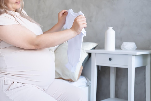 happy pregnant woman with big belly holding baby clothes in hands at home. motherhood concept