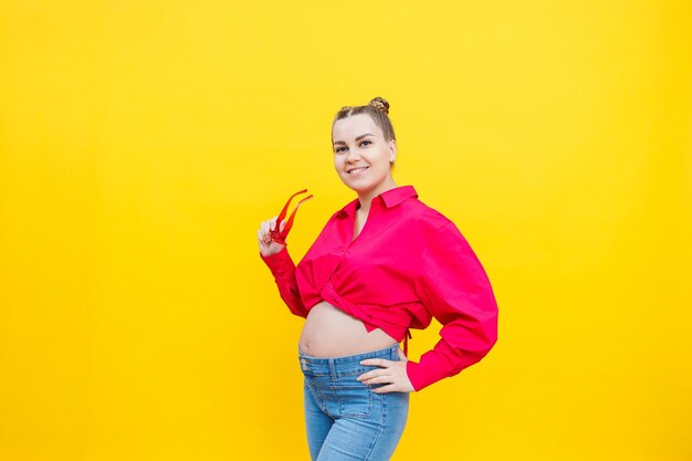 Happy pregnant woman with a smile on her face Cheerful pretty pregnant woman in a pink shirt and pink glasses on a yellow background Young bright pregnant woman