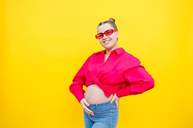 Happy pregnant woman with a smile on her face Cheerful pretty pregnant woman in a pink shirt and pink glasses on a yellow background Young bright pregnant woman