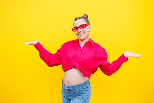 Happy pregnant woman with a smile on her face Cheerful pretty pregnant woman in a pink shirt and pink glasses on a yellow background Young bright pregnant woman