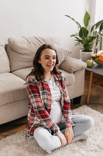 Happy pregnant woman woman in red plaid shirt and jeans laughing and sitting on carpet Brunette lady in white tee chilling out in living room