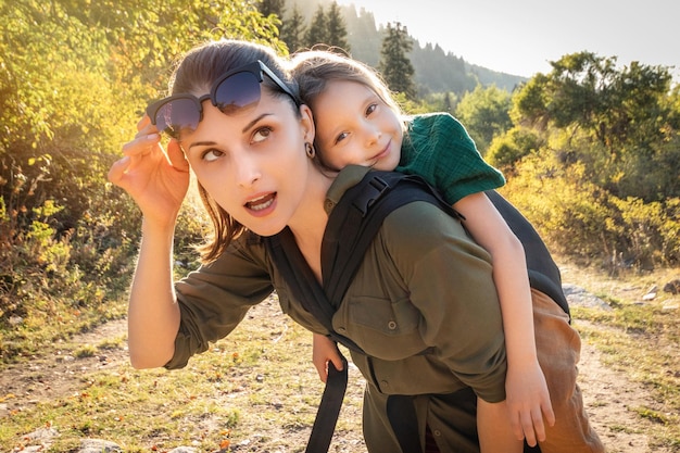 Happy preschool kid girl in babywearing sling at back her mother in hiking outdoor