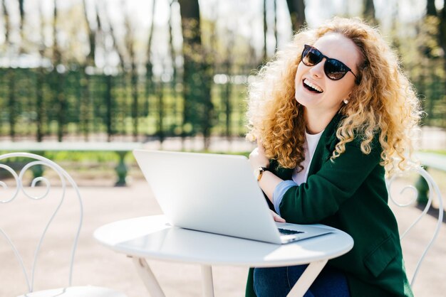 Happy pretty blonde woman with curly hair wearing sunglasses and green jacket having rest in park sitting at white desk using her laptop communicating with friends online People emotions concept