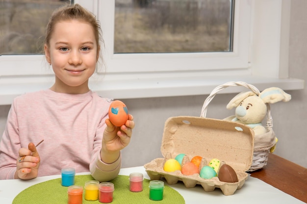 Happy pretty girl sitting at a table with paints and an easter bunny shows a painted easter egg