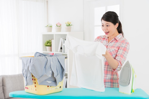 happy pretty woman looking at white clothing surprised clean and standing in front of ironing board doing housekeeping at home in living room.