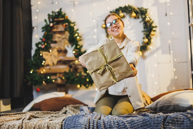 Happy pretty woman with fair hair in cozy clothes with a New Year's gift in her hands is sitting on the bed near the Christmas tree New Year's atmosphere at home