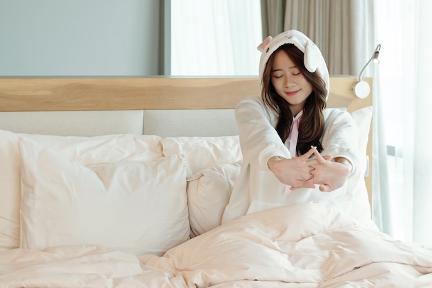 Happy pretty young Asian woman in pajamas sitting on bed and tsretching after waking up early in the morning