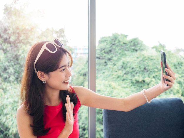 Happy pretty young Asian woman in pink casual shirts waving hand in good mood video call on mobile smartphone screen Closeup smiling young female greet her friends or family with relaxing at home