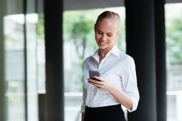 Happy pretty young businesswoman standing and using cell phone outdoors