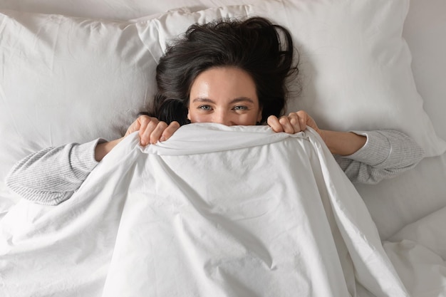 Happy pretty young european female sleeps on white bed covers face with blanket wakes up enjoys good morning