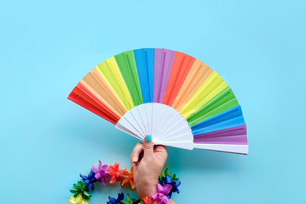 Happy Pride Month. Simple, minimal LGBT pride flag decor. Hand with fake nails in colors of the rainbow holds rainbow colored fan and flower garland. Flat lay, top view on mint green background..