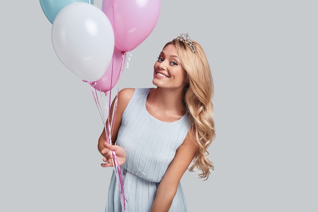 Happy princess. Attractive young woman holding balloons and looking at camera with smile while standing against grey background