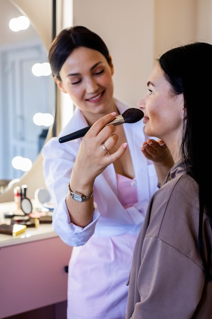 Happy professional makeup artist applying cosmetics lip pencil to woman client working beauty salon