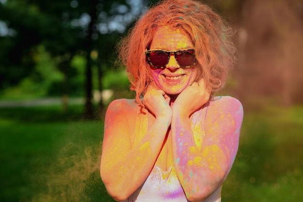 Happy redhaired woman wearing white t shirt posing covered with colorful dry Holi paint at the park