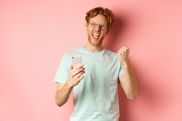 Happy redhead guy in glasses and t-shirt winning online prize, shouting yes with joy and satisfaction, holding smartphone and making fist pump, pink background