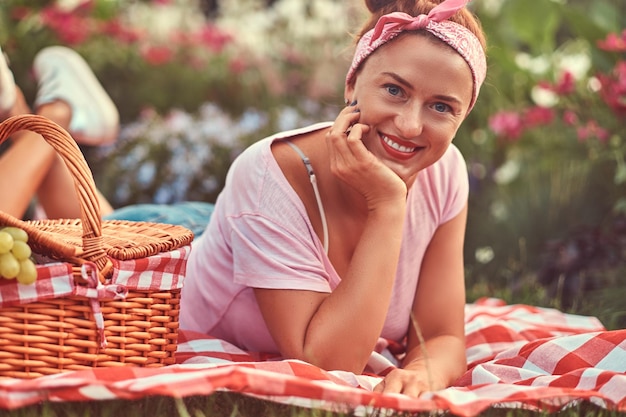 Happy redhead middle age female in casual clothes with a headband enjoying during picnic outdoors while lying on a blanket in the park.