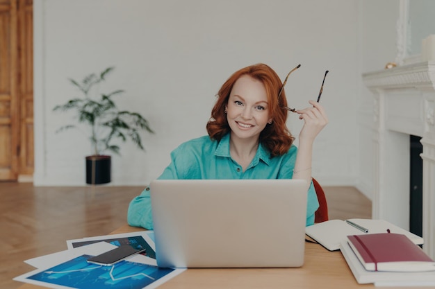 Happy redhead woman in coworking space works on laptop holds spectacles browses web communicates online Blue shirt paper documents