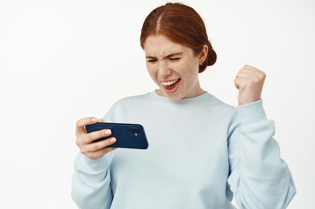 Happy redhead woman winning on mobile phone, playing video game on cellphone or watching video in internet, chanting, raising fist up and say yeah, white background