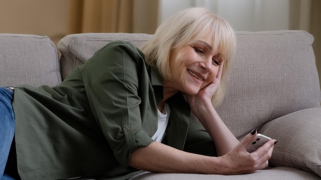 Happy relaxed Caucasian old senior mature woman holding smart phone looking at cellphone laughing