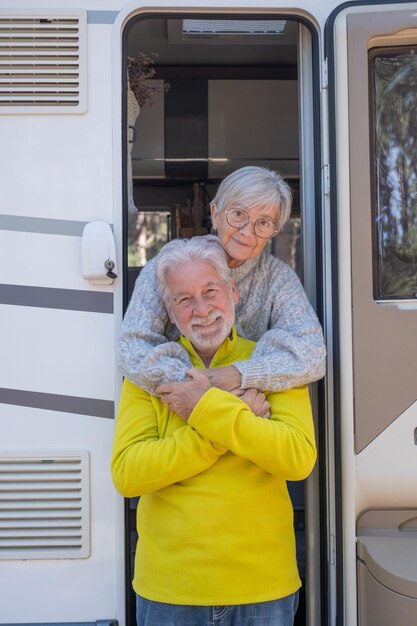 Happy relaxed caucasian senior couple on the door of their camper van motor home embrace looking at camera Smiling attractive elderly people enjoying freedom vacation travel in the forest