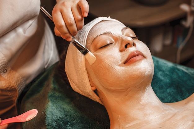 Happy relaxed woman leaning on a spa bed having a skin care white mask smiling in a wellness spa center while traveling.