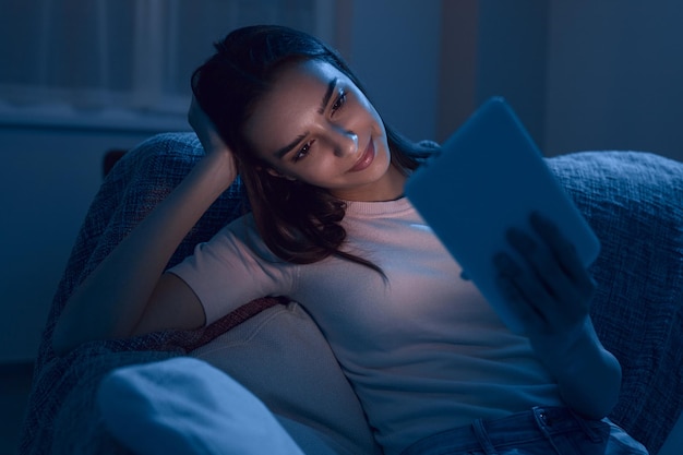 Happy relaxed young female sitting on sofa and reading e book on digital tablet while spending evening at home