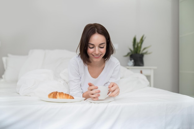 Happy relaxed young woman drinking coffee eating pastry in bed