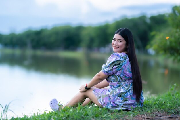 Happy Relaxing Portrait asian woman Wear purple dress wearing smartwatch while sitting on green grass lawn beside a reservoir at the city park outdoors