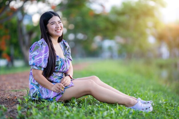 Happy Relaxing Portrait asian woman Wear purple dress wearing smartwatch while sitting on green grass lawn beside a reservoir at the city park outdoors