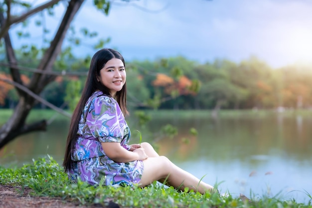 Happy Relaxing Portrait asian woman Wear purple dress wearing smartwatch while sitting on green grass lawn beside a reservoir at the city park outdoors