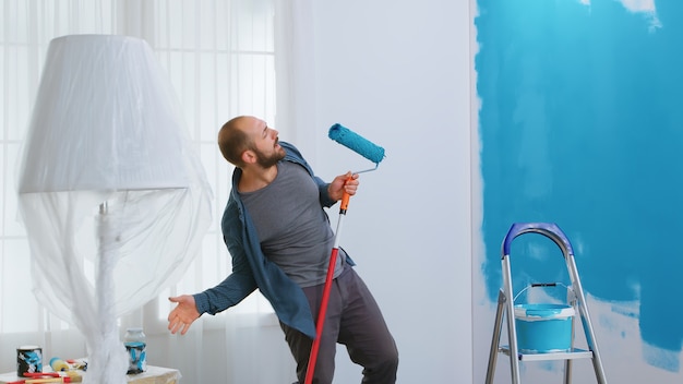 Happy repairman singing on roller brush during home renovation. Painting with blue paint. Apartment redecoration and home construction while renovating and improving. Repair and decorating.