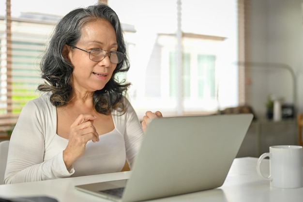 A happy retired Asian woman using her laptop at her home workspace