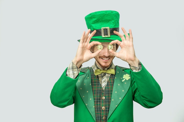 Happy rich young man in green suit hold two golden coins in front of eyes. He smile and look happy. Guy wear green saint Patrick's costume. Isolated on grey .