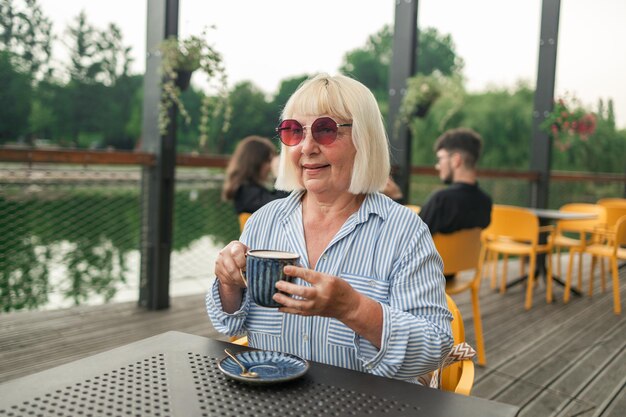 Photo happy s senior woman with stylish clothes and sunglasses drinking coffee at street cafe elderly old