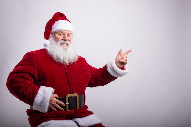 Happy Santa Claus, sat with hand in belt pointing a side in white wall