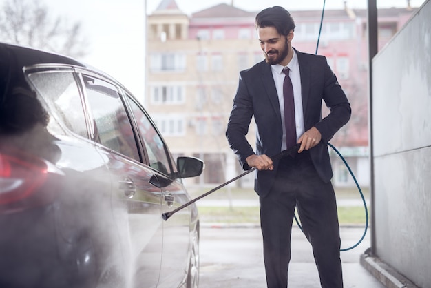 Happy satisfied elegant stylish young focused man in a suit washing the car with a water gun on self-service washing car station.