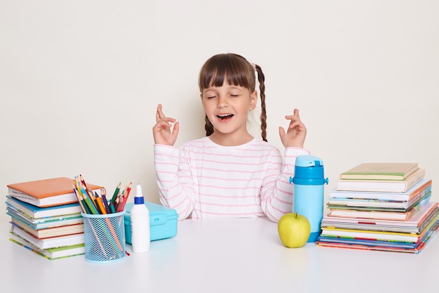 Happy satisfied hopeful little schoolgirl with pigtails wearing white shirt sitting at the desk with closed eyes crossed fingers making desire posing surrounded with books and other school supplies