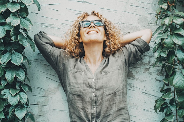 Happy and satisfied woman smiling and looking in the air with green nature ivy on both side Portrait of joyful female people standing agains an outdoor grey wall Concept of happiness One person