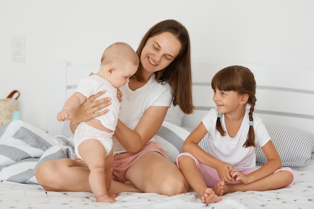 Happy satisfied woman wearing white shirt and short sitting on bed with her daughters in bedroom expressing optimistic emotions mother holding toddler baby and smiling family together