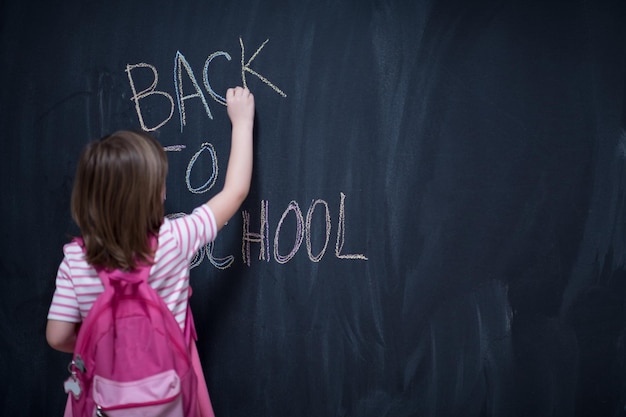 Happy school girl child with backpack writing  back to school on black chalkboard