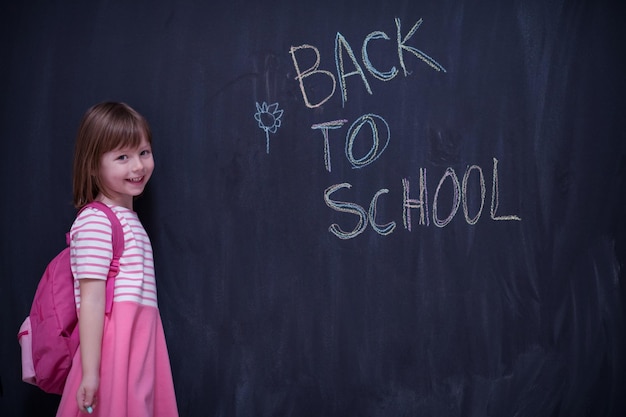 Photo happy school girl child with backpack writing  back to school on black chalkboard