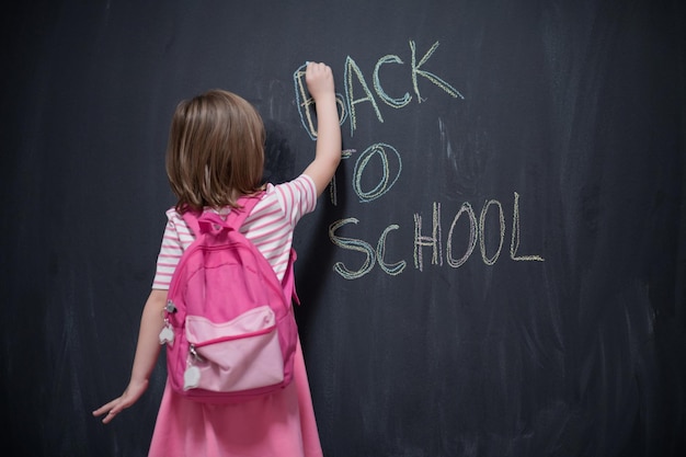 Happy school girl child with backpack writing  back to school on black chalkboard