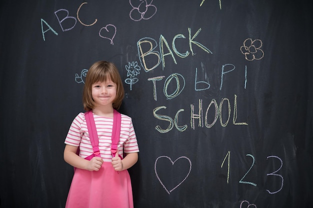 Photo happy school girl child with backpack writing  back to school on black chalkboard