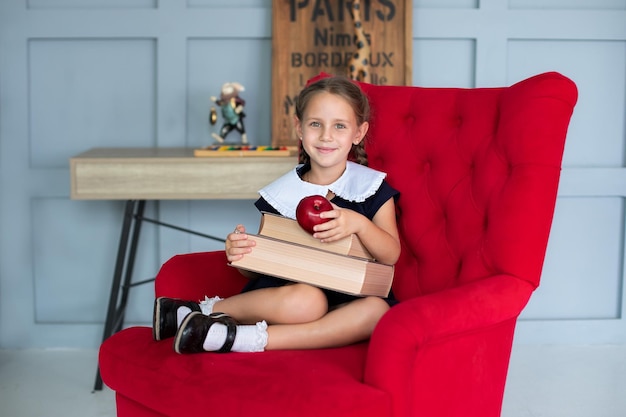 Happy School Girl in dress sit on chair and holds a stack of books in classroom on school