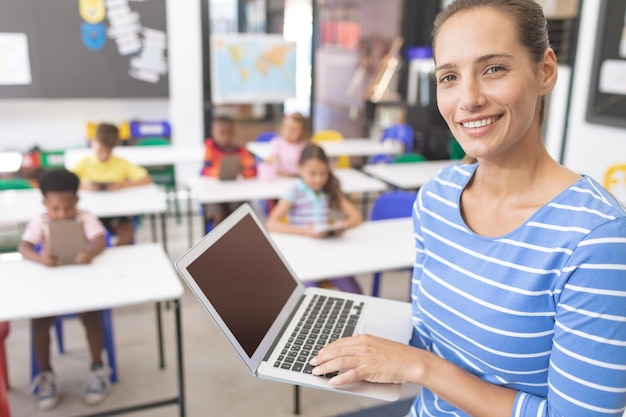 Happy schoolteacher standing in classroom with laptop