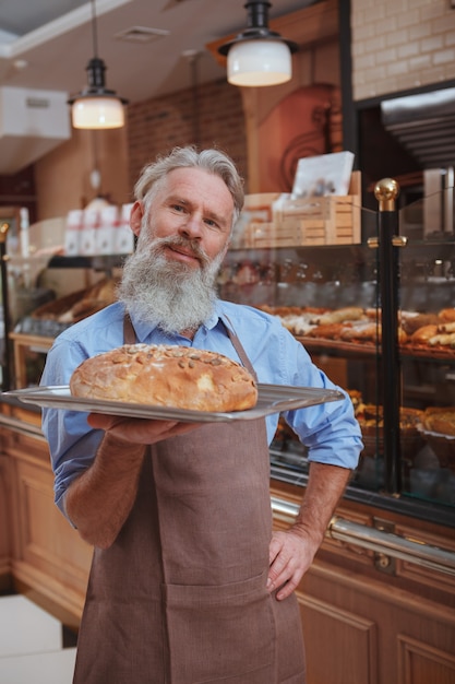 happy senior baker holding freshly baked bread on the tray smiling proudly to the camera