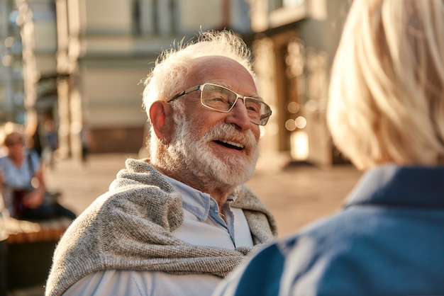 Happy senior bearded man in glasses looking at his wife and smiling while sitting on the bench